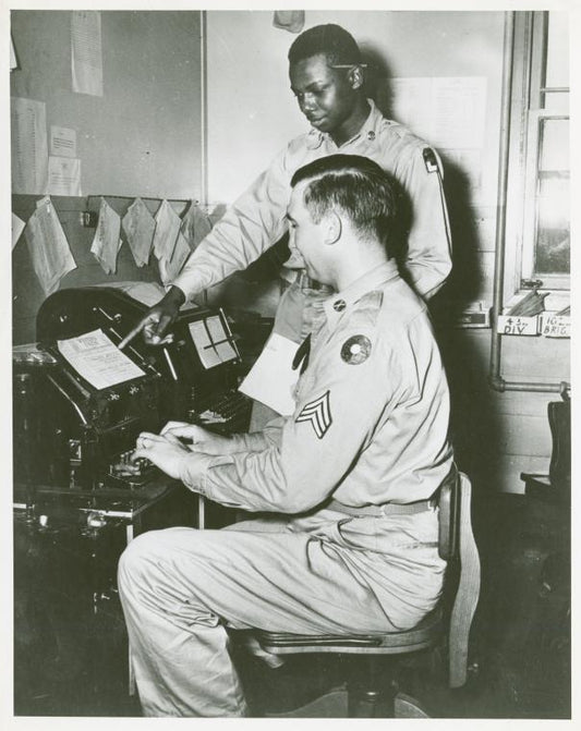 Corporal Parkins D. Peck, chief teletype operator, seated at the teletype machine while African American Private James Lee stands behind him and points to a Western Union telegram form, Camp Edwards, Kentucky